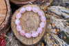 Pale pink bracelet with polished oval beads and cloudy white markings, arranged in a loop on a rustic wood slice surrounded by dried grasses, viewed from above.