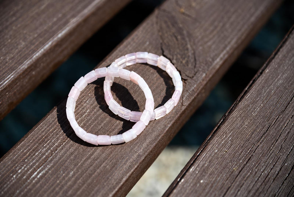 Two overlapping pale pink bracelets with square-shaped beads resting on dark wooden slats, viewed from above in sunlight with strong shadows.