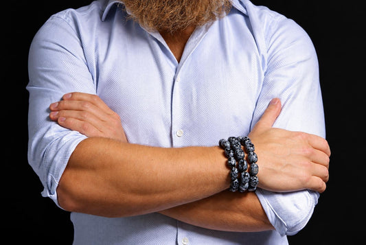 Three stacked bracelets of rounded black beads with grey mottled markings on a person's wrist, with crossed arms and a pale blue rolled-sleeve shirt against a dark background.