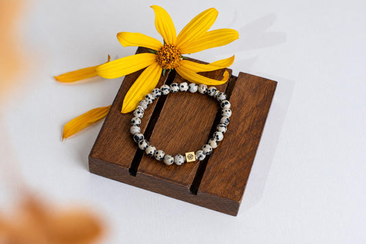 A bracelet with black and white spotted beads and a gold-toned square charm rests on a dark wooden stand beside a yellow flower.