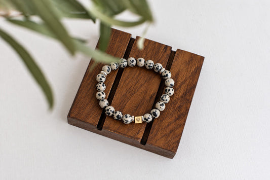 A bracelet with white and black spotted beads, a gold-coloured square charm, and silver-toned spacers rests on a dark wooden surface.