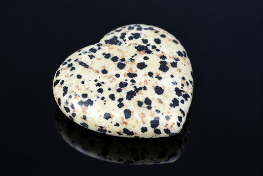 A heart-shaped stone with a cream background and black spots, resting on a dark reflective surface.