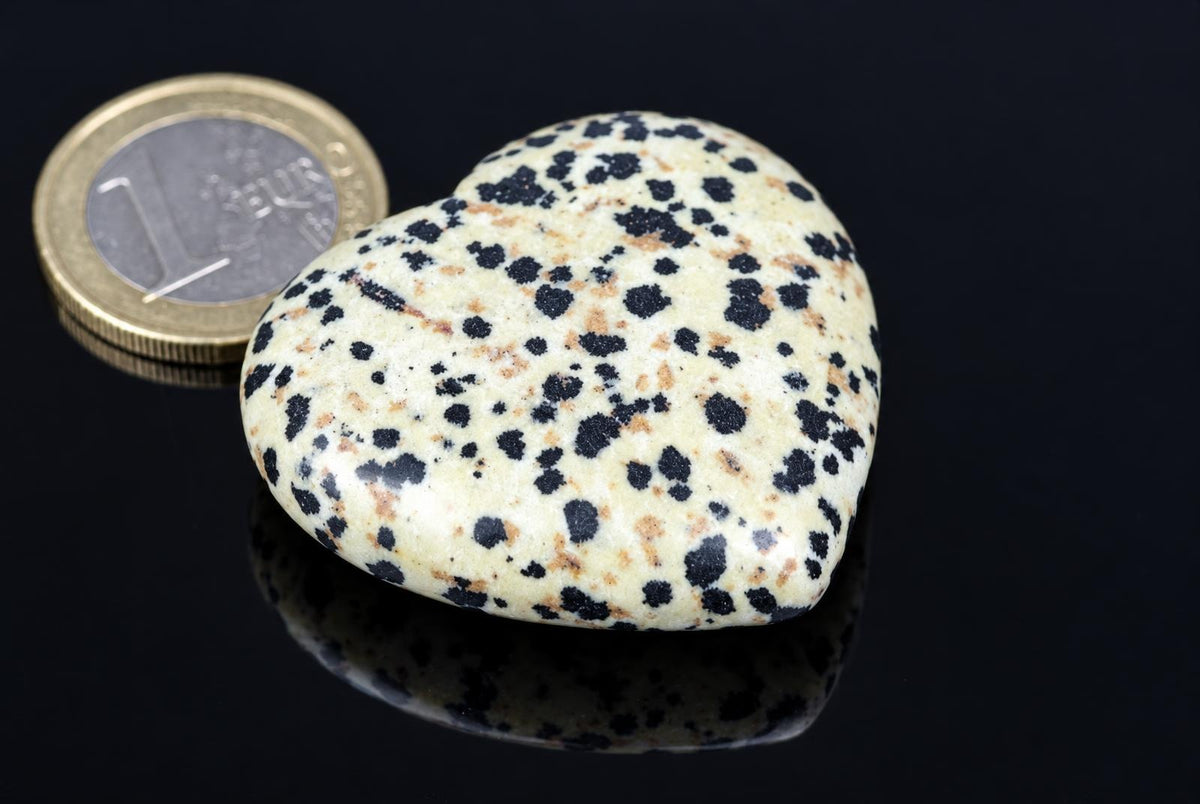 A heart-shaped stone with a cream background and black spots sits on a dark surface next to a coin.
