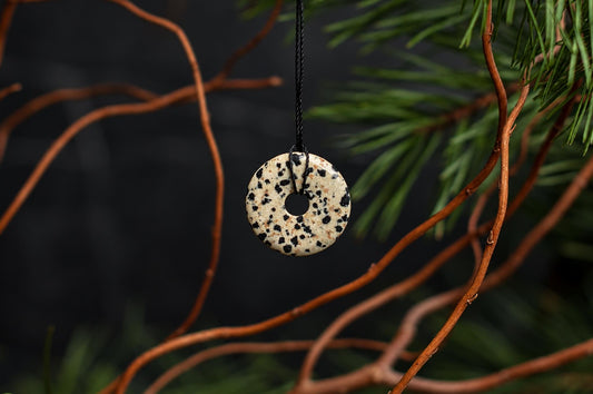 A cream-coloured round pendant with black spots and a central hole hangs from a black cord among green needles.
