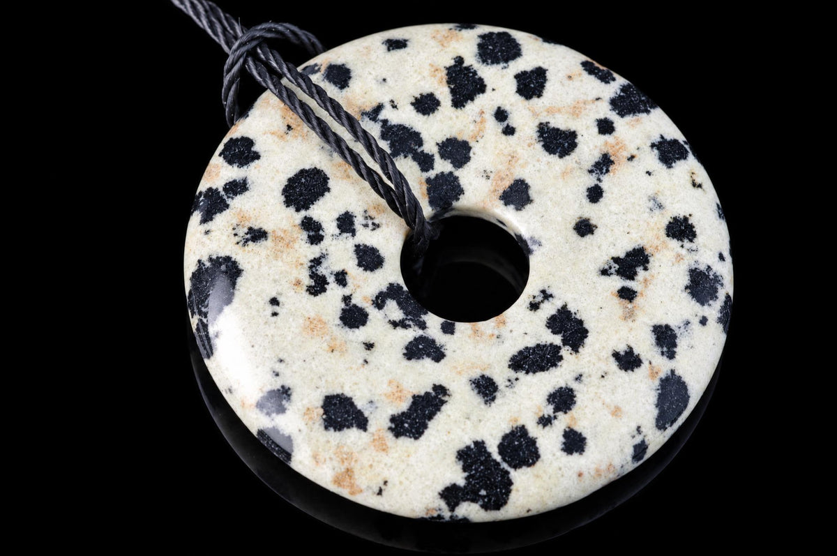 A round, donut-shaped pendant with a white base and black spots, featuring a central hole and a dark cord against a black background.