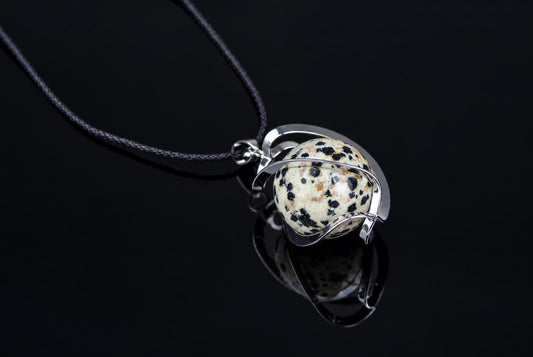 A silver-toned pendant with a spotted white and black spherical stone hangs from a dark cord against a dark, reflective surface.