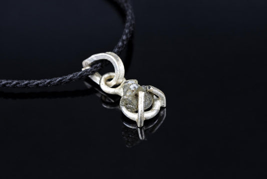 A silver-toned pendant with a rough, greyish stone set in an open loop design on a black cord against a dark surface.