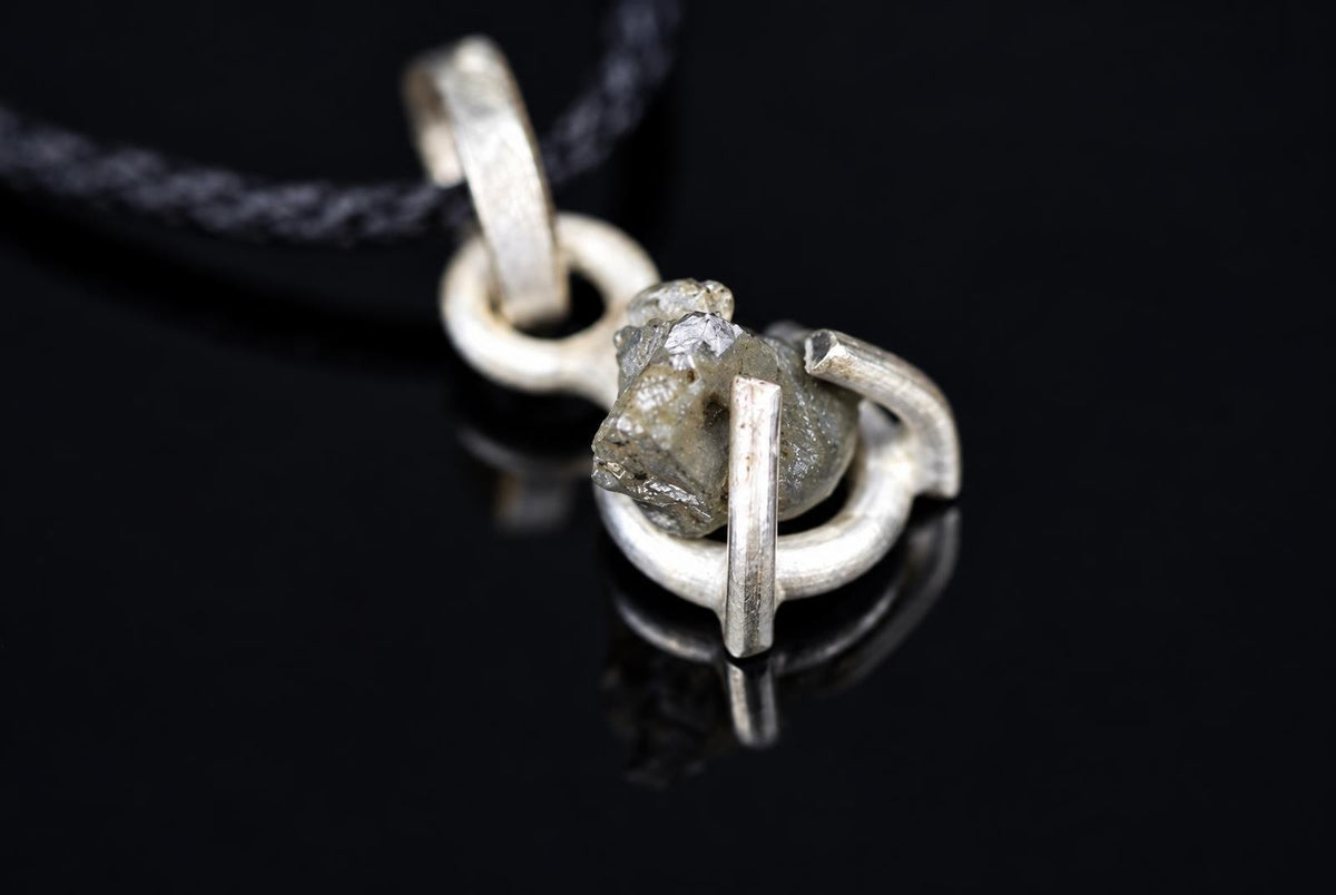 A silver-toned pendant with a rough, greyish stone held in a loop design on a black cord against a dark surface.