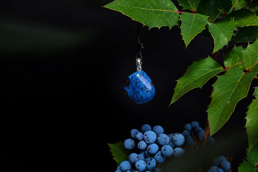 A blue dumortierite pendant with black speckles hangs from a silver bail on a dark cord, set against green leaves and berries.