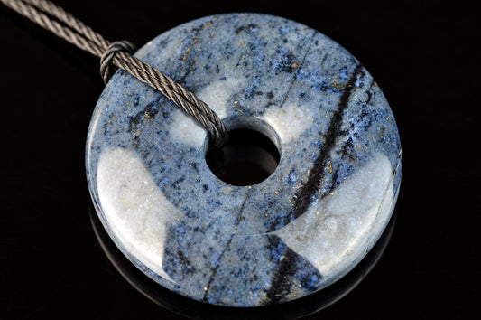 A polished, donut-shaped pendant with blue and white speckled stone on a dark surface, threaded with a grey cord.