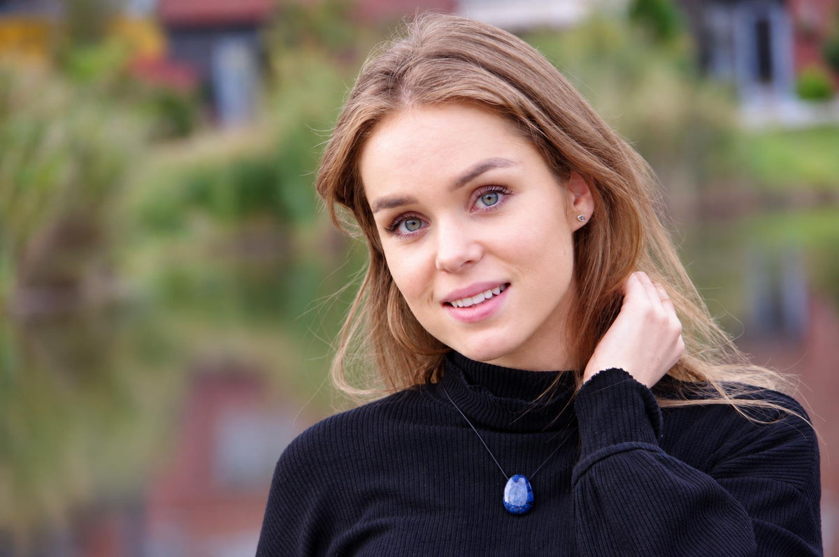 A woman wears a black turtleneck sweater with a dark blue, teardrop-shaped pendant on a thin cord against a blurred outdoor background.