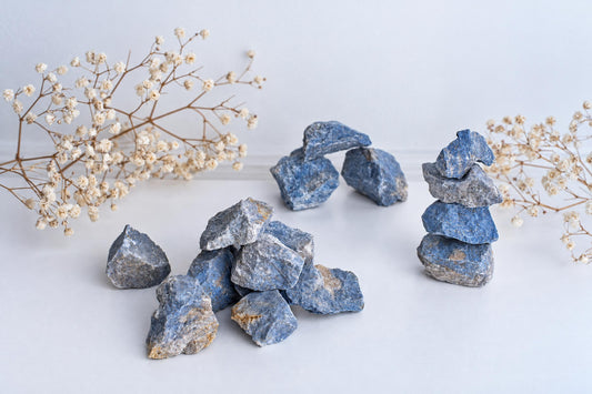 Blue and grey Dumortierite raw stones arranged in piles on a pale surface with dried flowers.