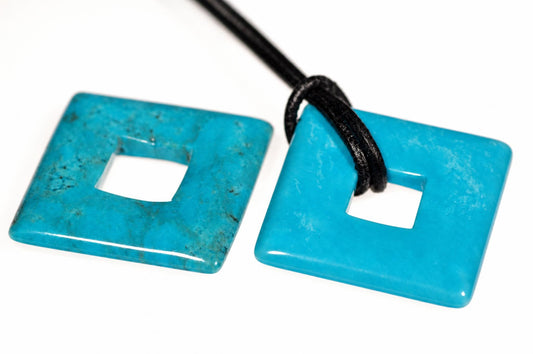 Blue-green square pendants with central square openings lie on a white surface, one threaded onto a black cord.