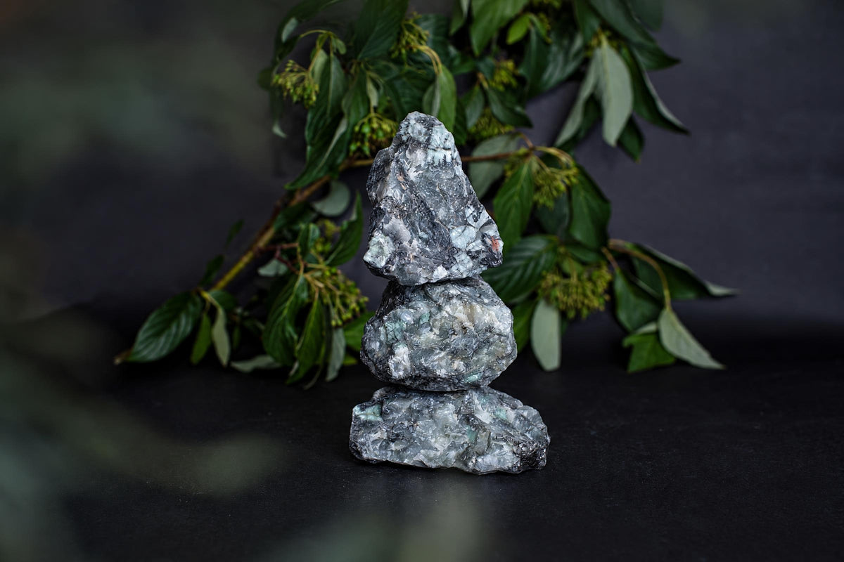 Three stacked rough emerald crystals on a dark surface with green foliage in the background.