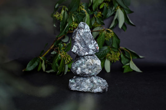 Three stacked rough emerald crystals on a dark surface with green foliage in the background.