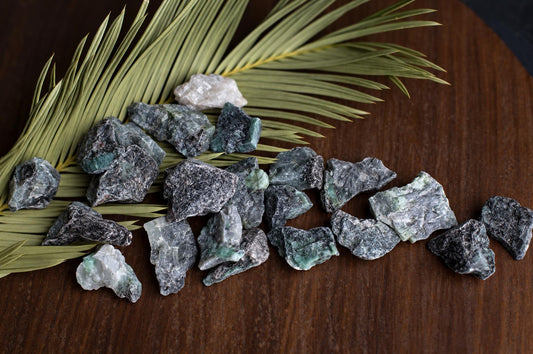 A pile of raw emerald rough crystals with dark grey and green hues rests on a wooden surface beside a dried palm leaf.