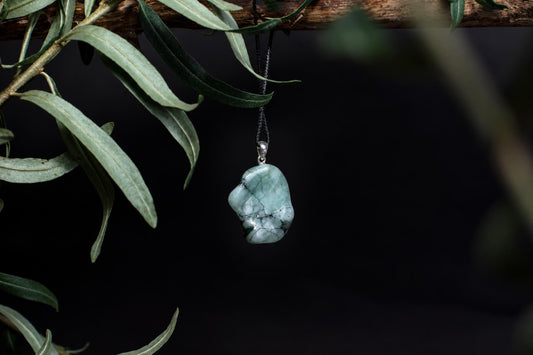 A polished, light green pendant with dark veins hangs from a black cord on a branch against a dark background.