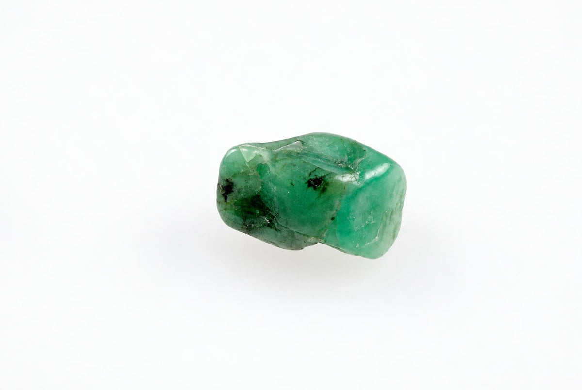 A polished green emerald stone with dark inclusions rests on a white surface.