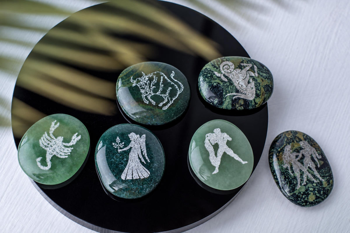 Six polished green stones with silver-toned engravings of zodiac symbols and figures rest on a dark circular surface.
