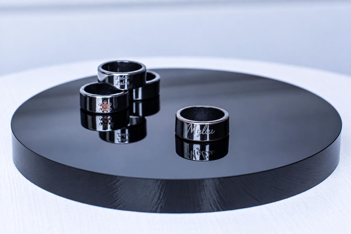 Black rings with engraved lettering and symbols sit on a round black reflective display against a pale background.