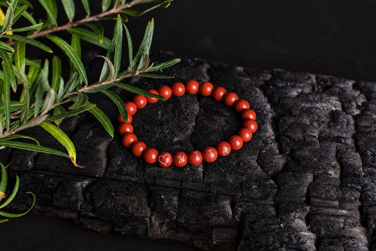 A red beaded bracelet with white engraved symbols rests on a dark, textured wooden surface beside green foliage.