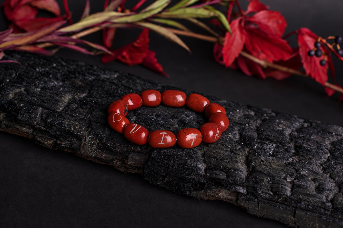 A red beaded bracelet with engraved letters and a heart rests on a dark wooden surface, surrounded by red leaves.