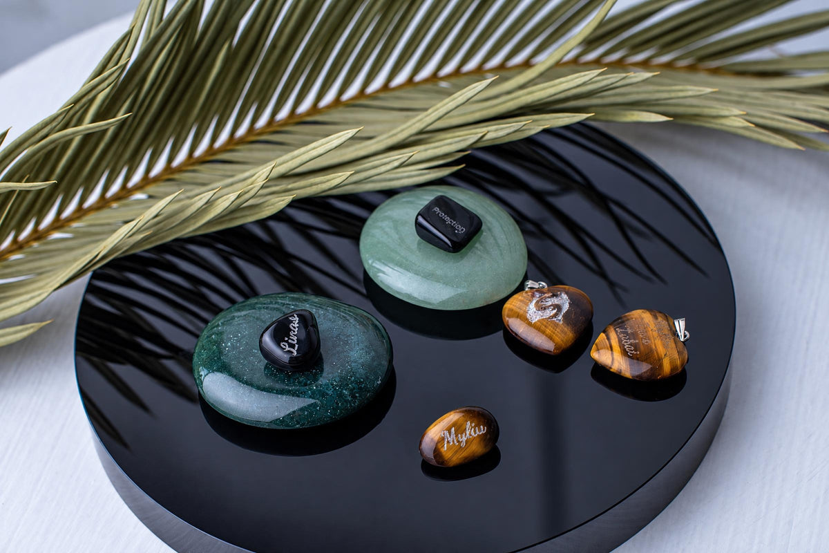 Small engraved dark stones rest on green circular stones beside brown polished stones on a round black tray beneath long green leaves.