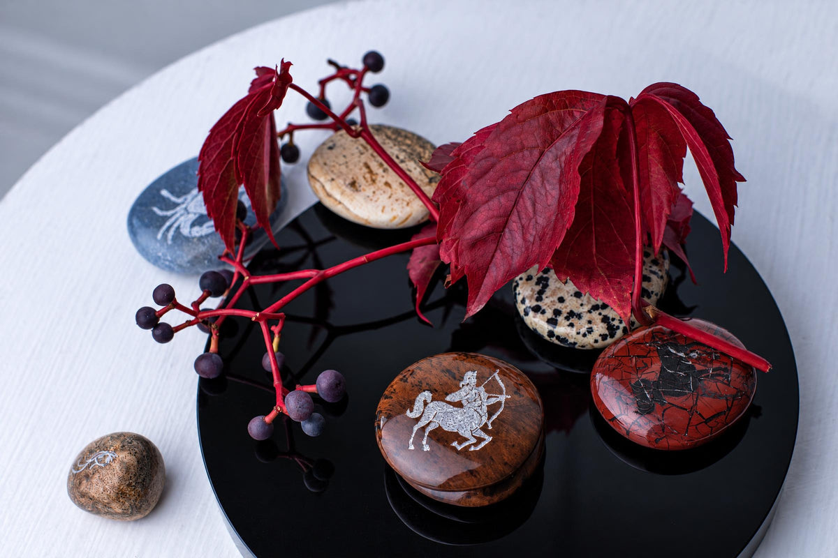 A black tray holds smooth, round stones with white engravings of a centaur and other figures, arranged with red leaves and berries on a pale surface.