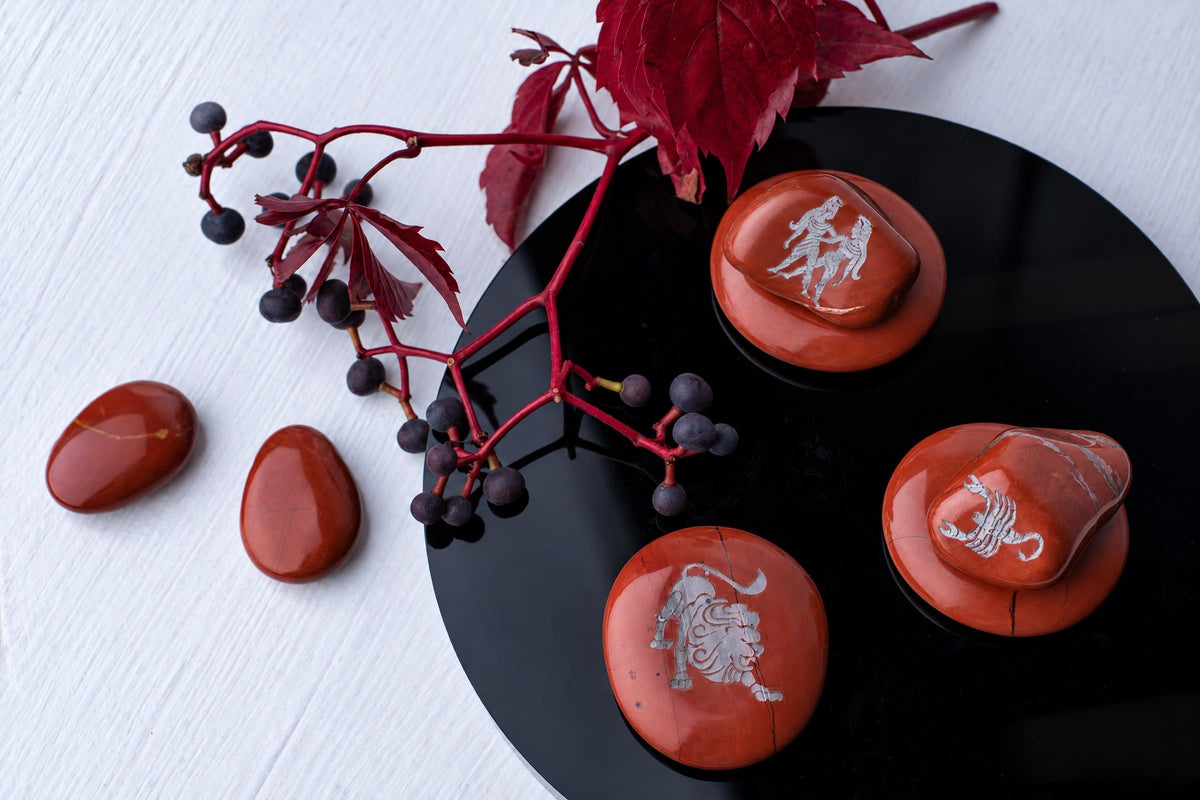 Red polished stones with silver-toned engravings of a lion, scorpion and figures rest on a black circular surface beside red leaves.