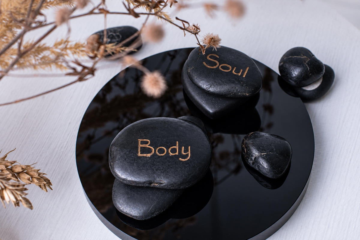 Smooth black stones engraved with words and small drawings sit on a glossy round black tray, with dried stems above a pale surface.