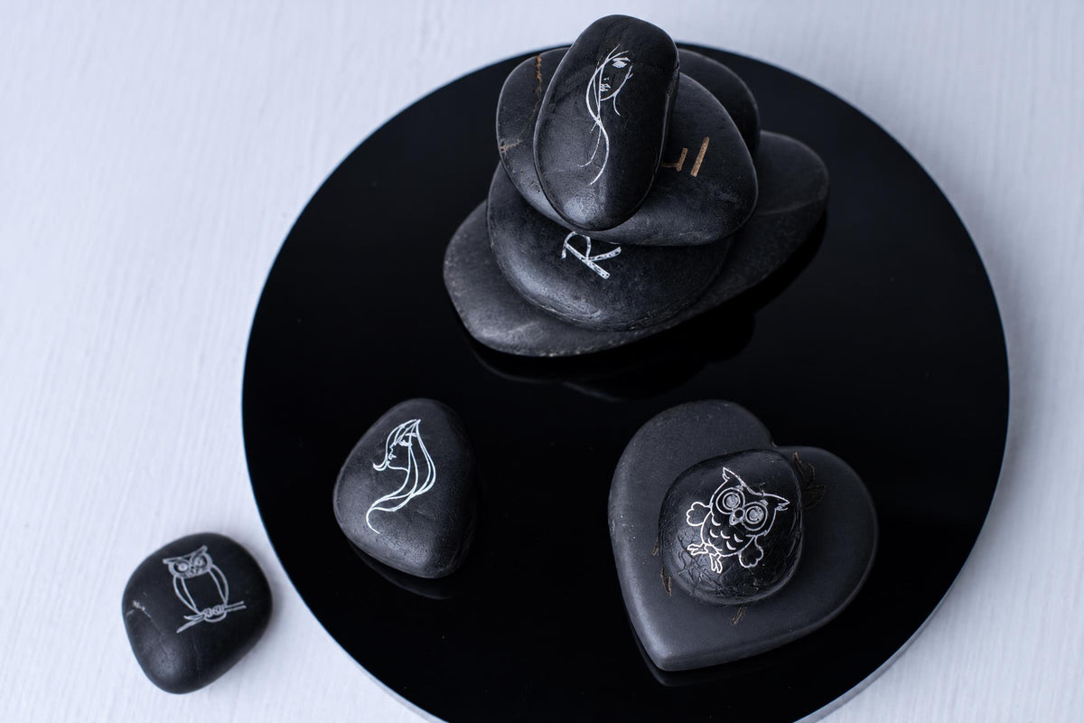 Black engraved stones with white designs, including owls and faces, arranged on a dark circular surface against a pale background.