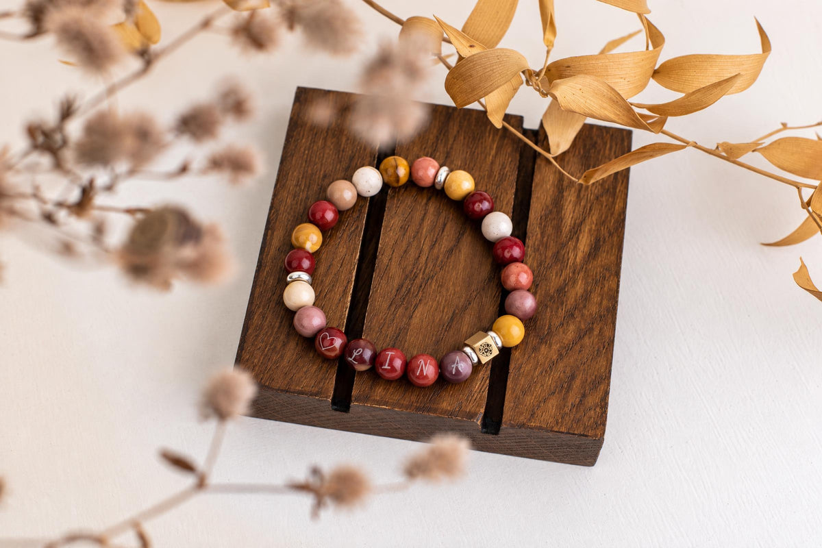 A beaded bracelet with red, yellow, white and pink beads sits on a dark wooden stand. Some beads are engraved with letters.