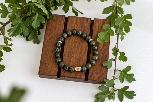 A bracelet with dark green speckled beads and silver-toned accents rests on a wooden surface, surrounded by green leaves.