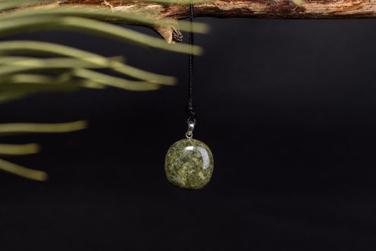 A round, polished green stone pendant hangs from a black cord on a dark surface.