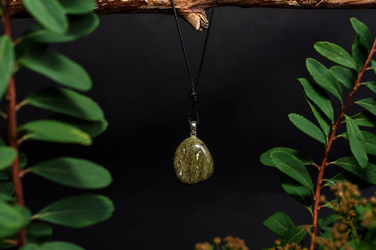A polished green stone pendant hangs from a black cord on a dark surface, framed by green leaves.