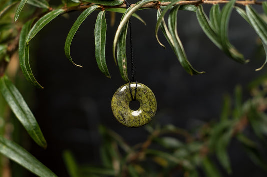 A polished green donut-shaped pendant hangs from a black cord on a leafy branch against a dark background.