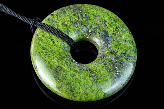 A polished, green epidote pendant shaped like a donut with a central hole, featuring dark speckles and light inclusions, on a black cord against a dark surface.