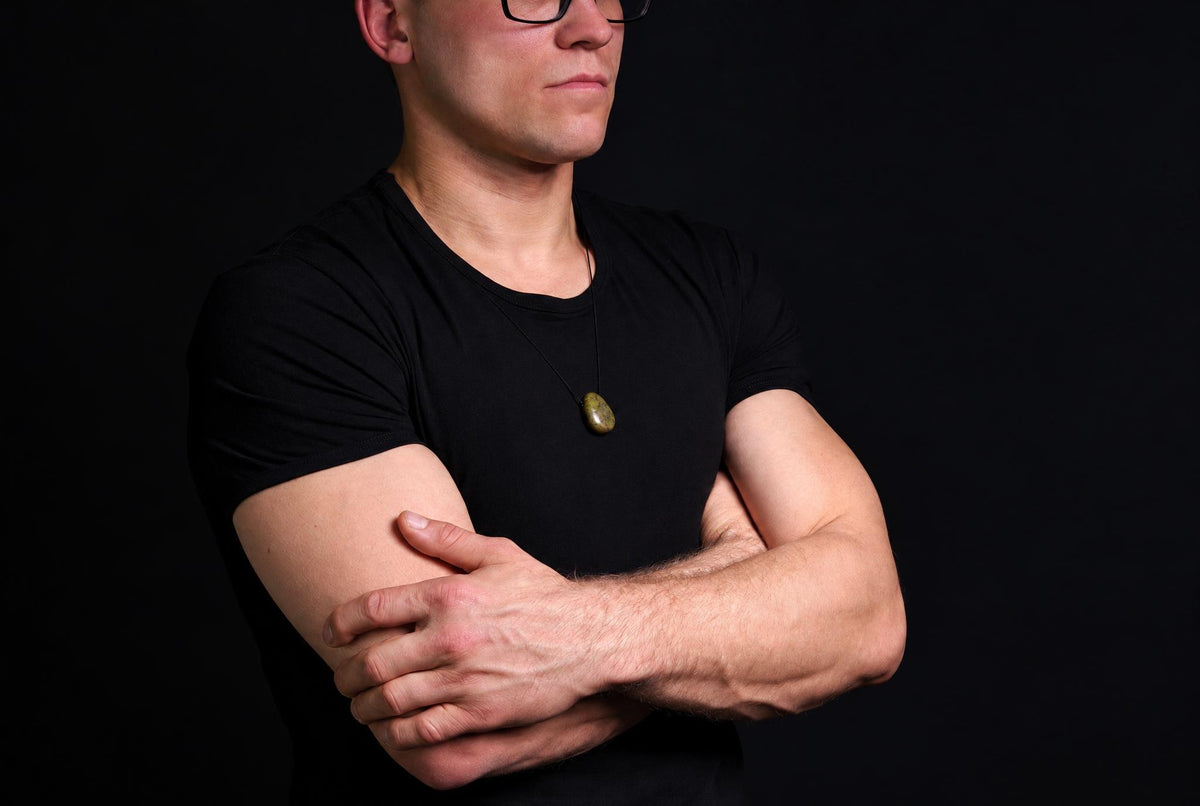 A man in a black t-shirt wears a dark green, oval-shaped pendant on a thin cord against a dark background.