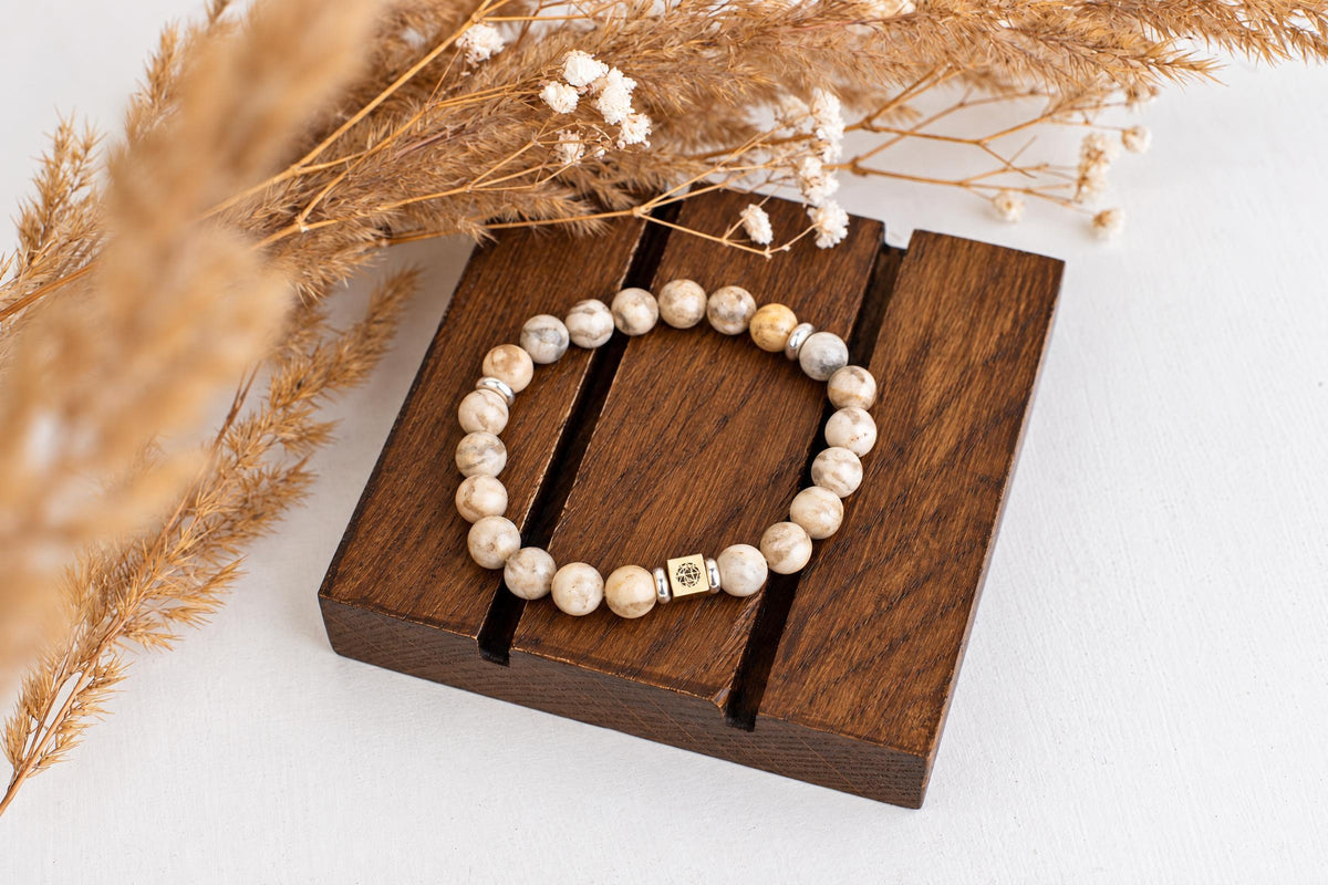 A beaded bracelet with cream and grey patterned stones, a gold-toned square charm, and silver spacers rests on a dark wooden block.