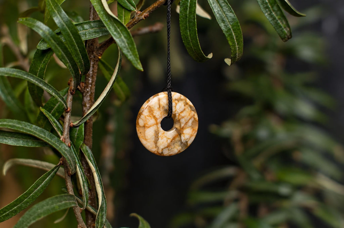 A polished, donut-shaped pendant with a swirling pattern of cream and brown hangs from a black cord on a leafy branch.