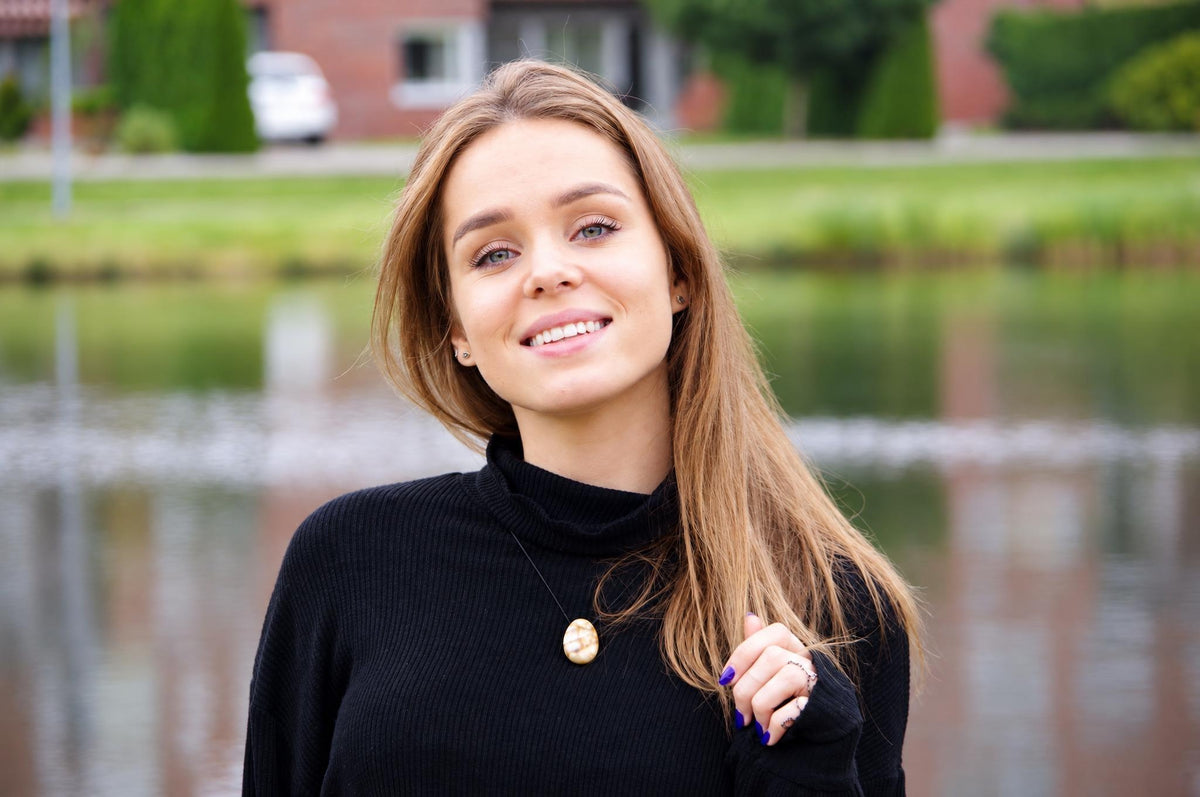 A woman wears a black turtleneck with a gold-coloured oval pendant necklace, standing outdoors near water.