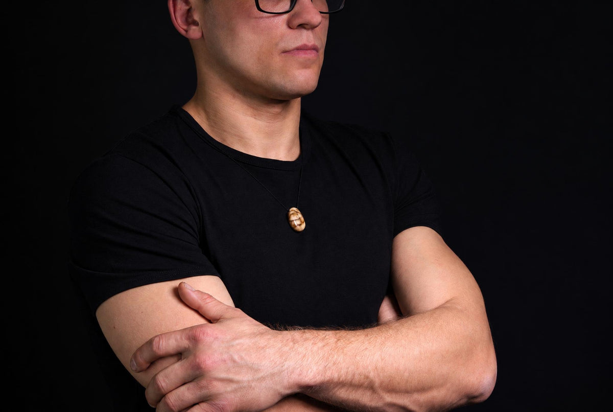 A man in a black t-shirt wears a pendant with a brown and cream patterned drop shape on a dark surface.