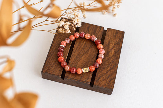 A bracelet of rounded reddish beads and small metallic spacers rests on a wooden board beside dried stems.