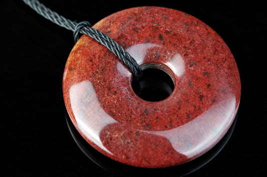 A polished, red and orange speckled donut-shaped pendant with a central hole, threaded on a dark grey cord against a black background.