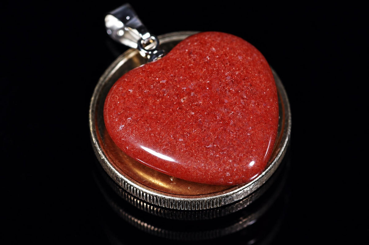A polished red heart-shaped stone pendant set in a silver-toned bezel with a bail, resting on a dark reflective surface.