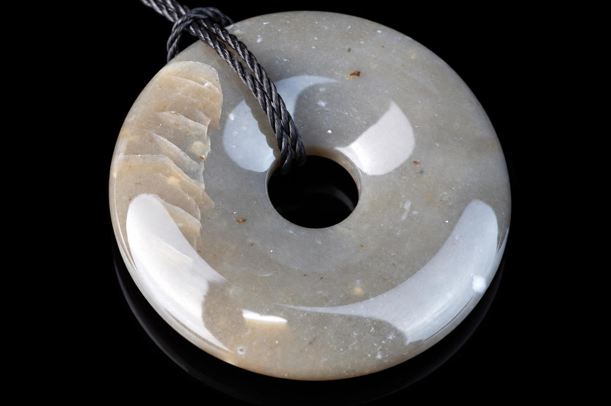 A polished, donut-shaped pendant with a central hole and a dark cord. It has a smooth surface in pale grey, beige, and white tones against a black background.