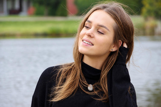 A woman with long brown hair wears a black turtleneck sweater and a light-colored oval pendant necklace, smiling outdoors near water.