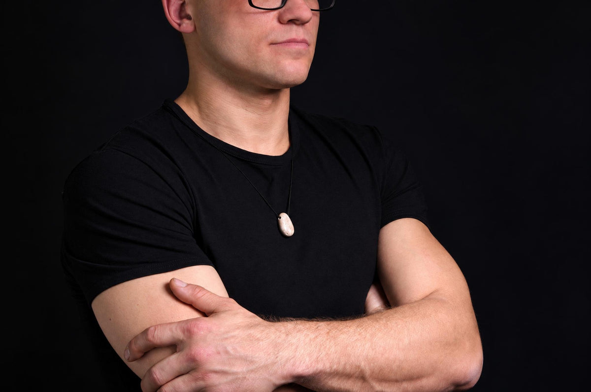 A man in a black t-shirt wears a pendant with an oval, light-coloured stone on a dark cord against a dark background.