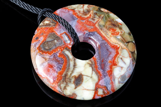 A polished, donut-shaped pendant with a central hole and a black cord. The stone displays swirling patterns of red, white, purple, and brown on a dark surface.
