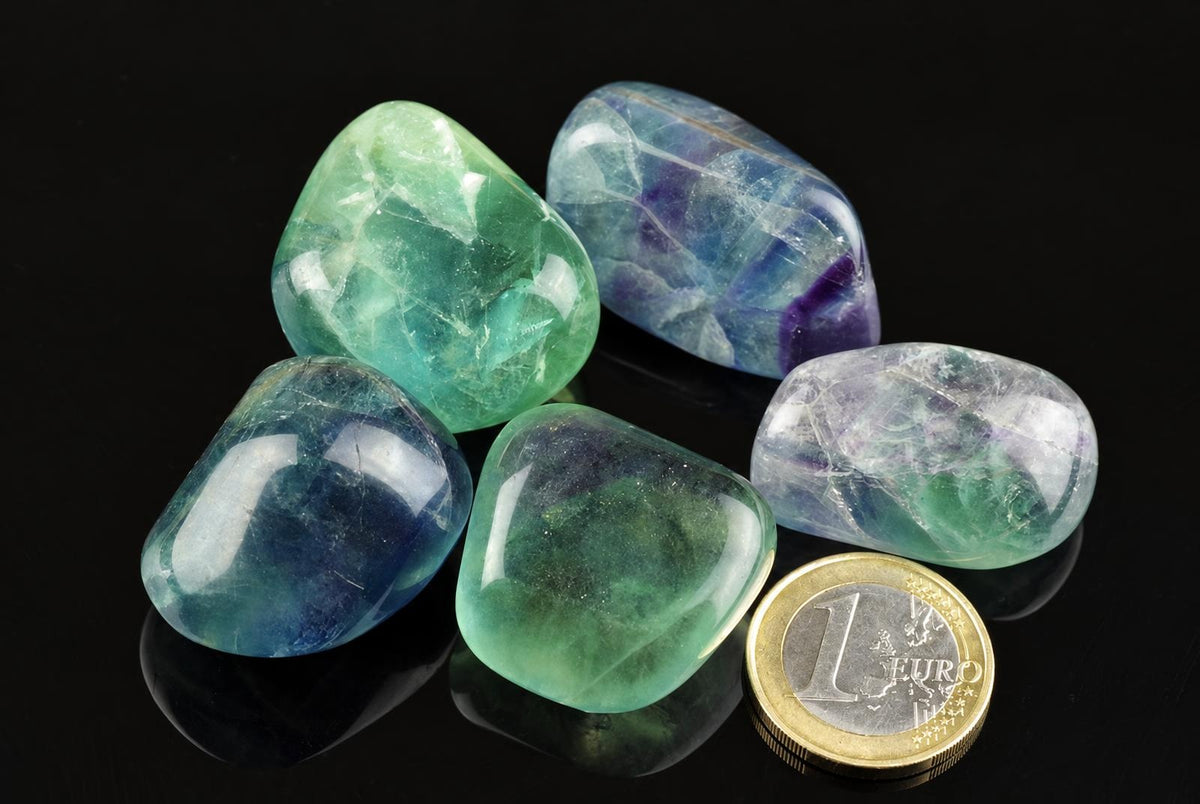 Five polished fluorite stones in green, blue and purple hues rest on a dark surface next to a one euro coin.